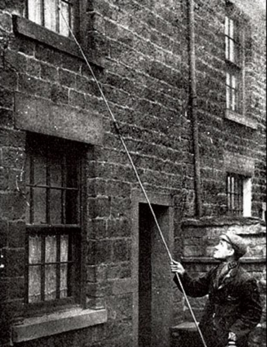 The Knocker-Up at work in Blackburn. He uses a long stick with a knob on the end to knock at bedroom windows and wake the resident ready for work.